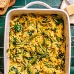 Baked orzo pasta with asparagus, spinach, and grated cheese in a white ceramic baking dish on a green tiled surface.