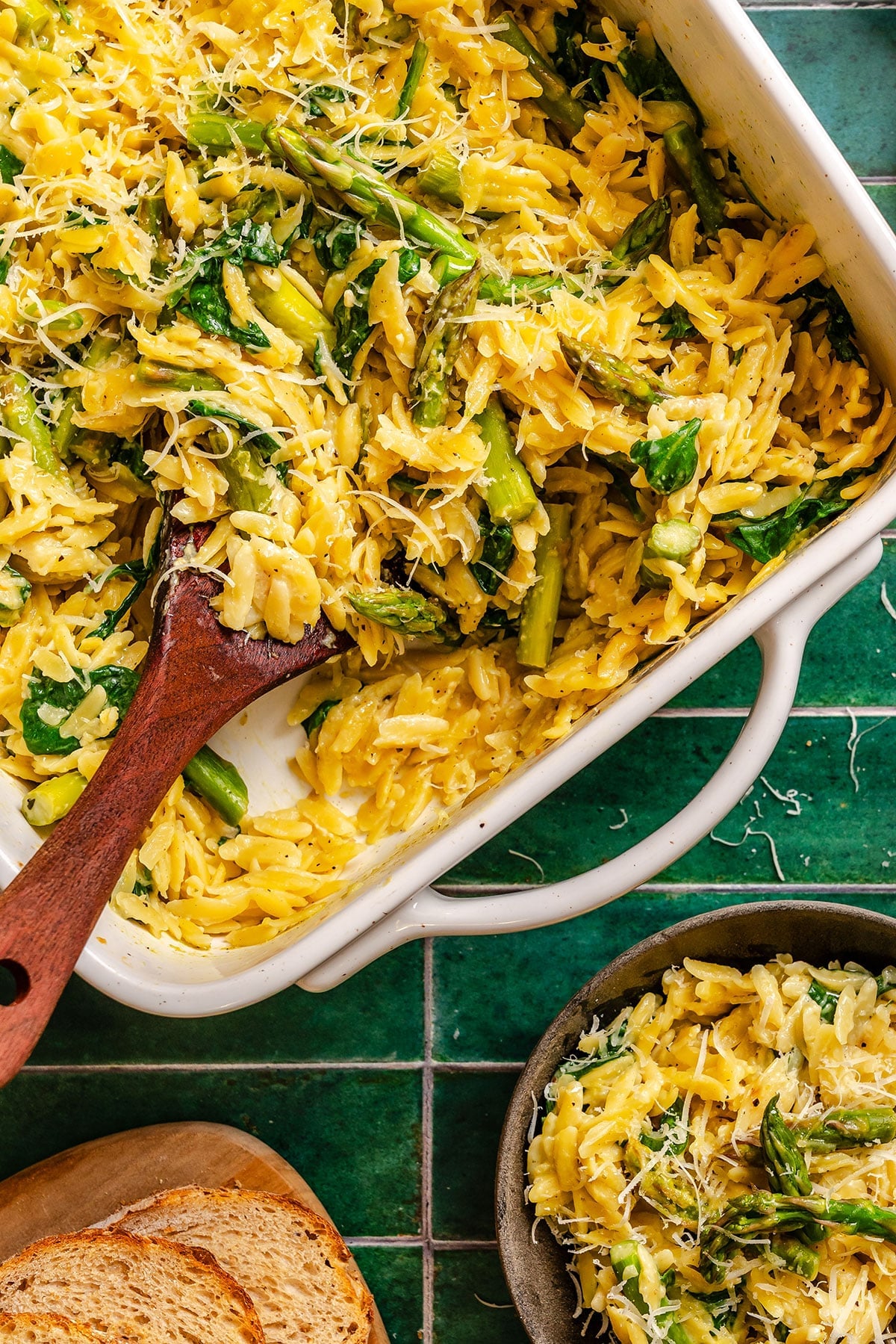 A white baking dish filled with orzo pasta, asparagus, spinach, and grated cheese sits on a green tiled surface next to a bowl of the same dish and slices of bread.