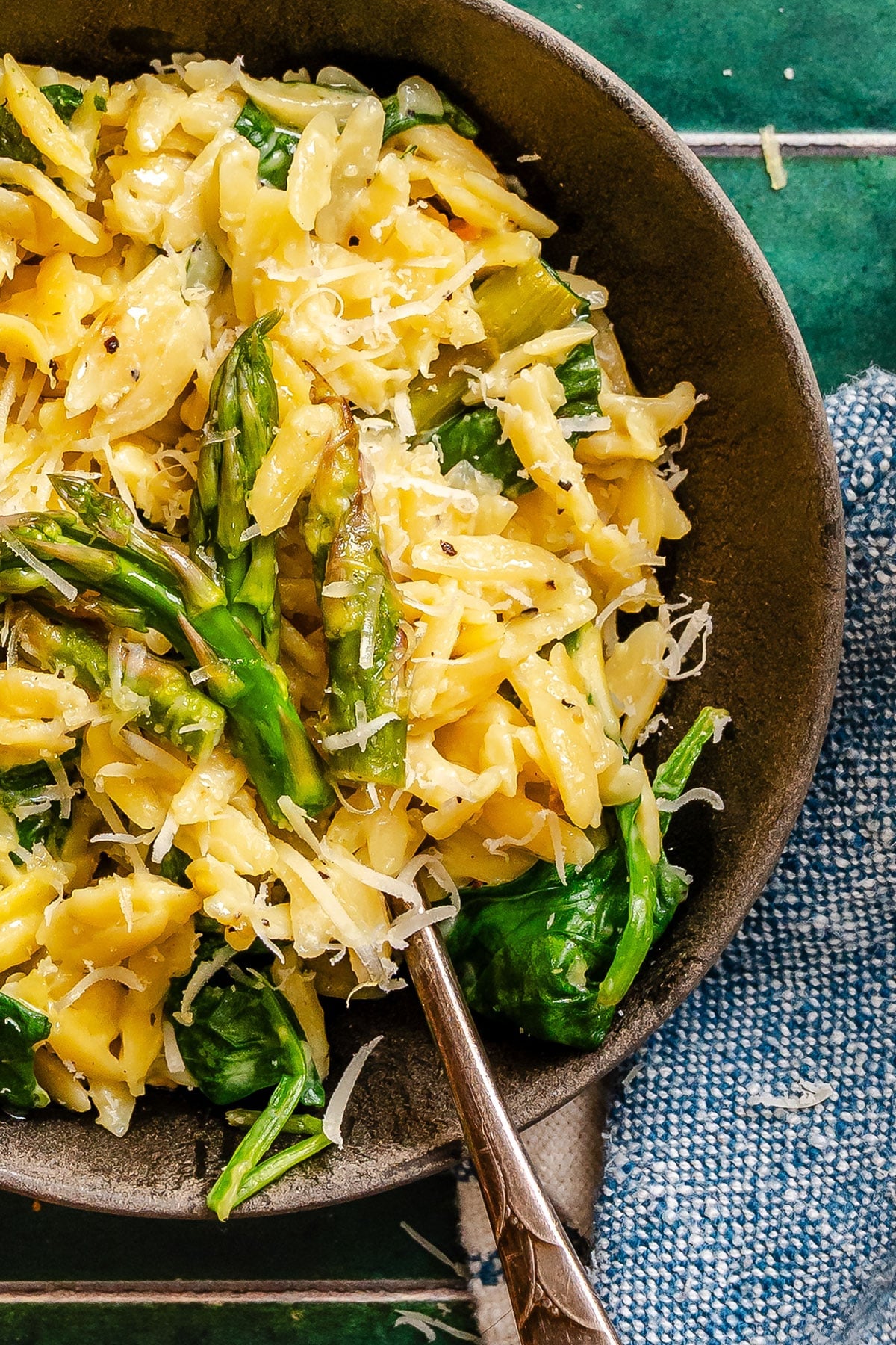 A bowl of orzo pasta mixed with asparagus, spinach, and grated cheese, with a fork resting in the dish on a green tile surface next to a blue textured napkin.