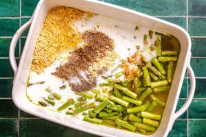A white baking dish filled with uncooked orzo, asparagus, broth, minced garlic, salt, pepper, and seasonings sits on a green tiled surface—perfect for an easy Dump and Bake Orzo Recipe.