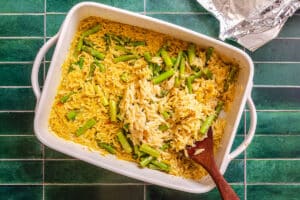 A white baking dish filled with orzo pasta and asparagus sits on a green tiled surface, with a wooden spoon resting inside and a piece of foil partially visible nearby.