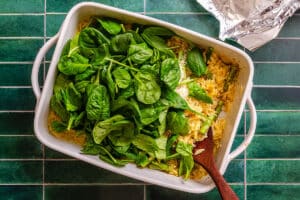 White baking dish with cooked pasta and fresh spinach leaves on top, partially mixed with a wooden spoon, on a green tiled surface.