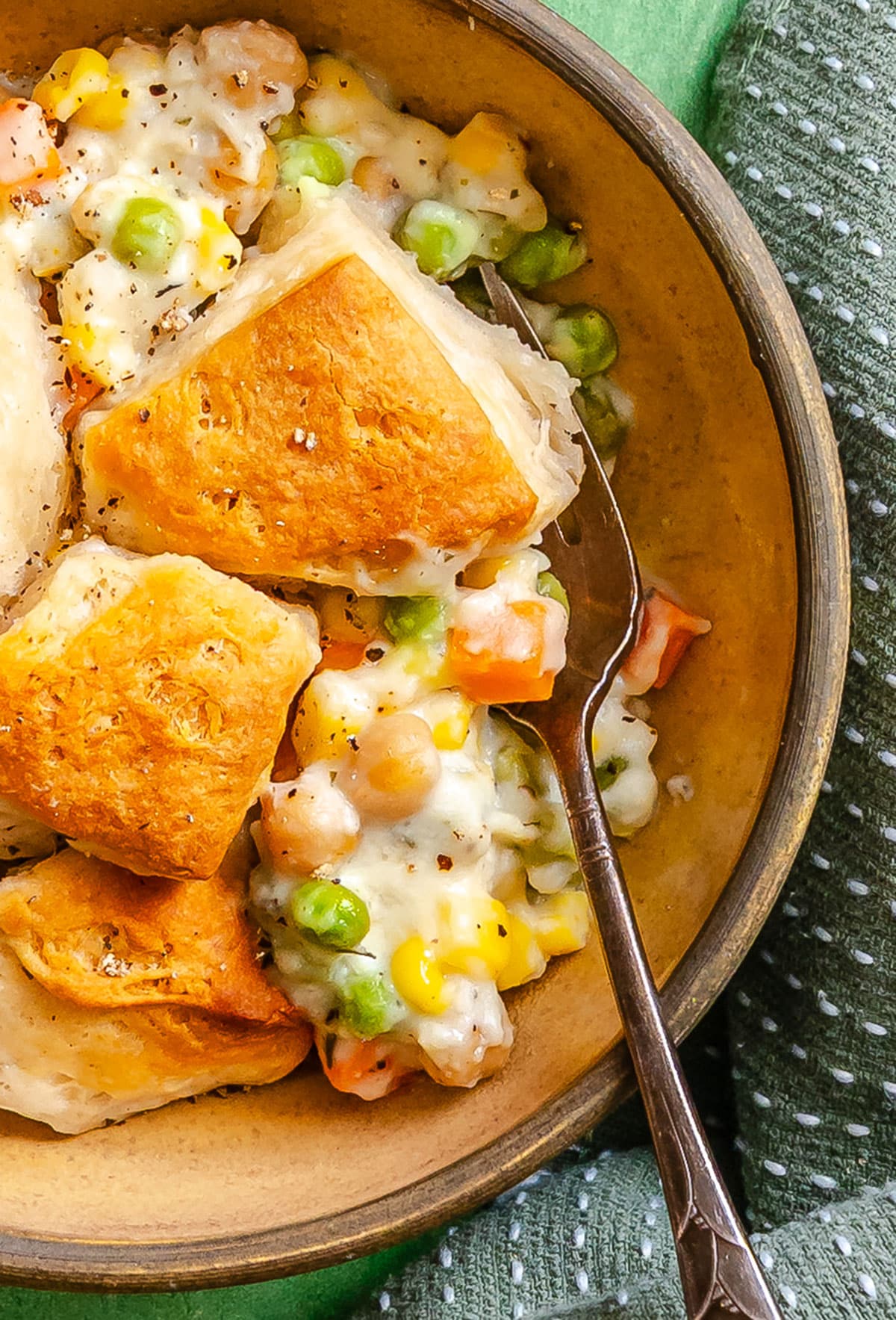 A bowl of biscuit-topped vegetable pot pie casserole with peas, corn, carrots, and creamy sauce, shown with a spoon on a green cloth—a comforting dump and bake classic.