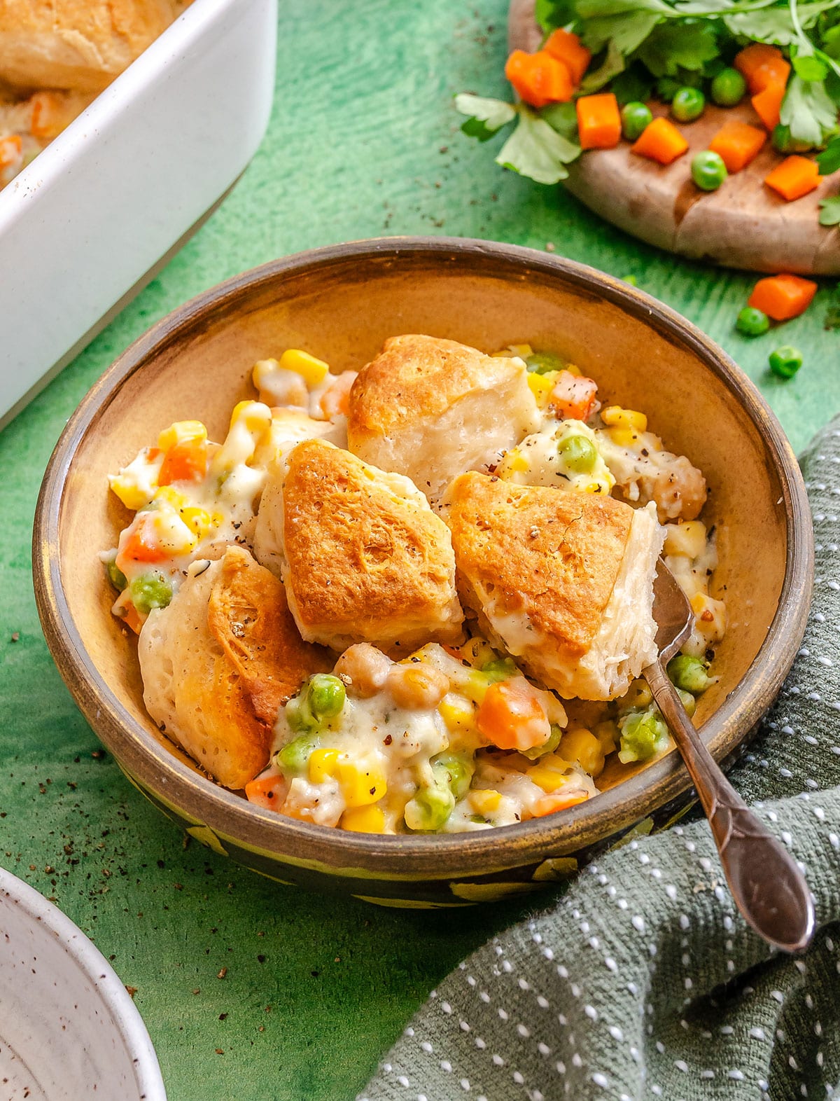 A bowl of vegetable pot pie casserole with carrots, peas, and corn in a creamy sauce, served with a spoon on a green surface, fresh vegetables in the background—a cozy dump and bake dish for any meal.