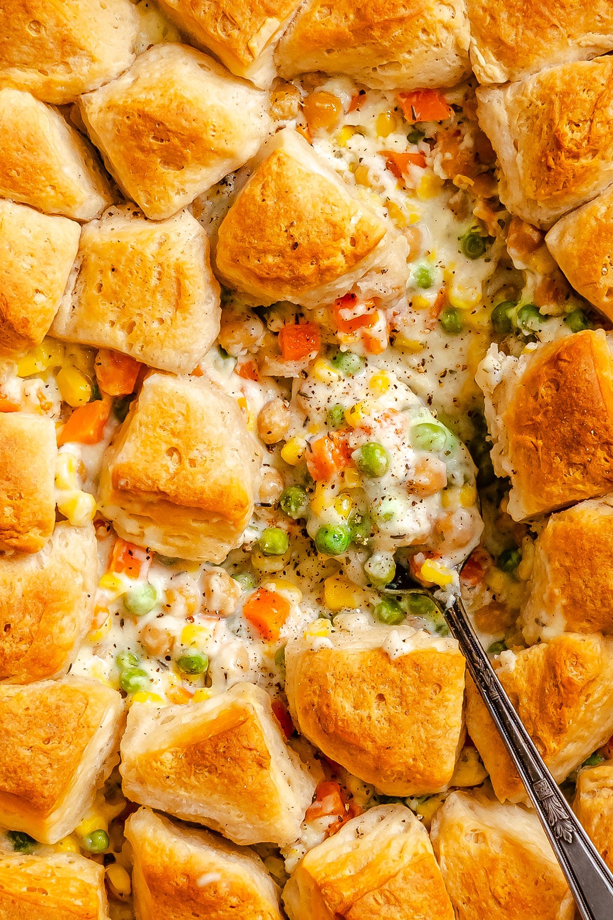 A close-up of a biscuit-topped Vegetable Pot Pie casserole with mixed vegetables and creamy sauce, as a spoon scoops out a hearty portion.