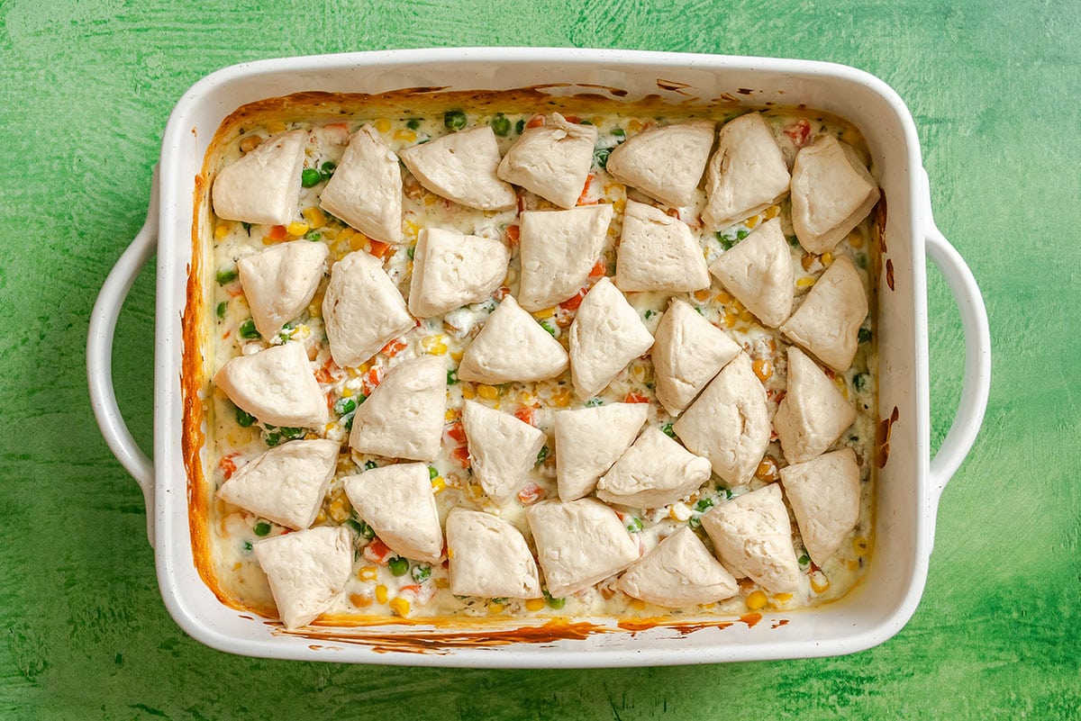 A casserole dish of creamy mixed vegetables, topped with unbaked biscuit pieces for a dump and bake vegetable pot pie, sits ready for baking on a green surface.