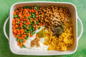 A white baking dish containing peas, diced carrots, chickpeas, corn, yogurt, and various seasonings arranged in separate sections on a green surface—perfect for a colorful Dump and Bake Vegetable Pot Pie casserole.