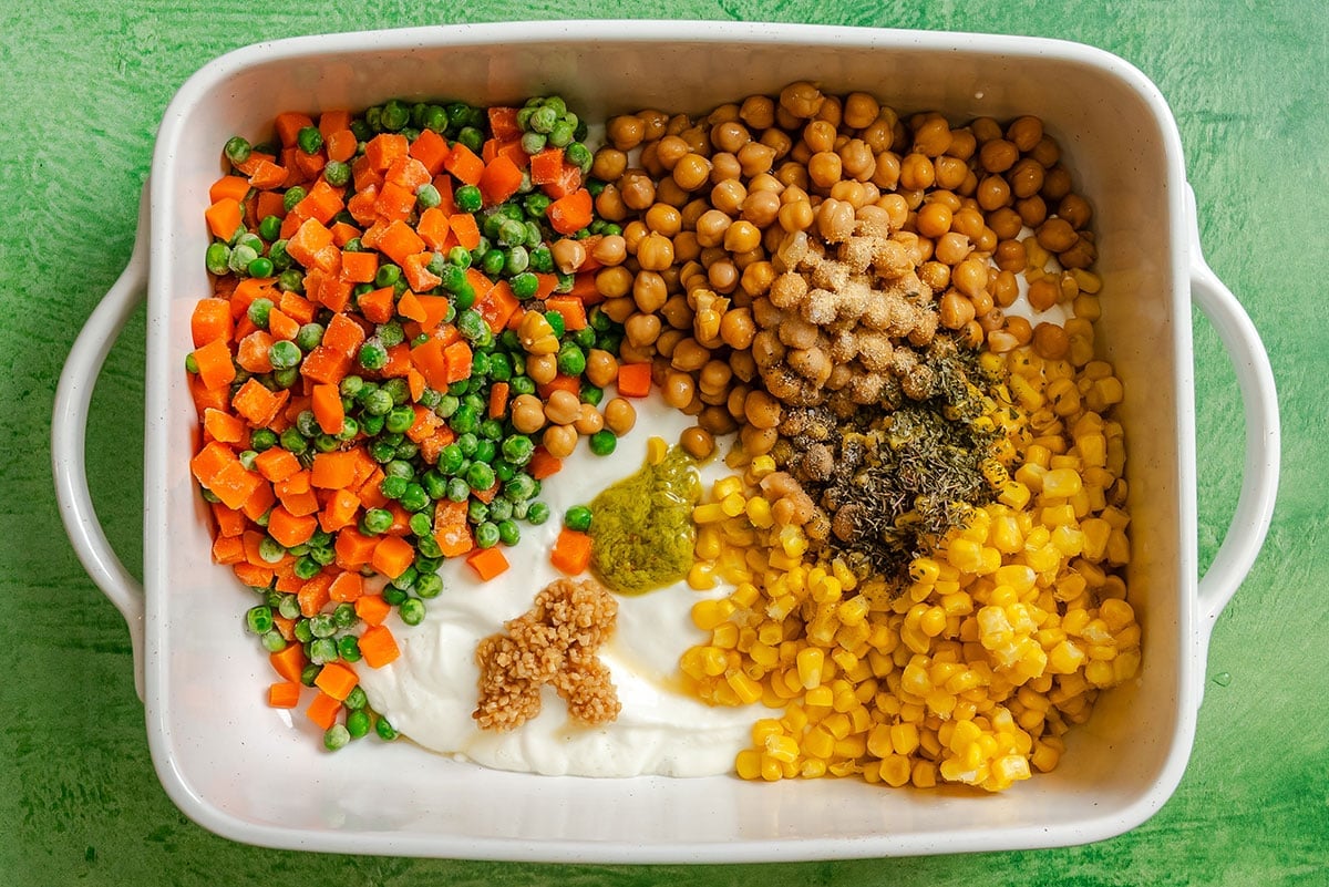 A white baking dish containing peas, diced carrots, chickpeas, corn, yogurt, and various seasonings arranged in separate sections on a green surface—perfect for a colorful Dump and Bake Vegetable Pot Pie casserole.