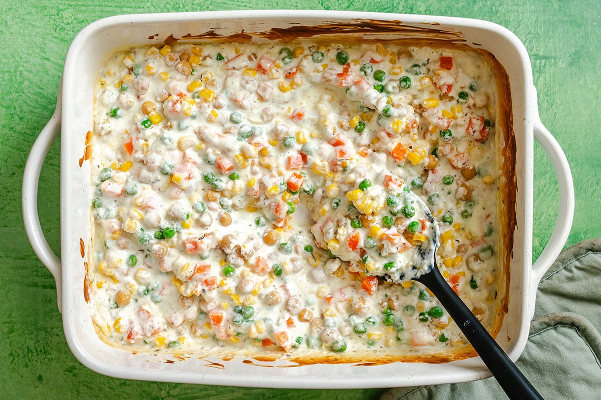 A white baking dish filled with creamy vegetable pot pie casserole, with a black serving spoon scooping out a portion—an easy dump and bake comfort meal.