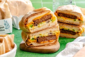 Two breakfast sandwiches with plant-based sausage, egg, cheese, mushrooms, and green peppers are stacked on a wooden board, wrapped in paper, with more sandwiches in the background.