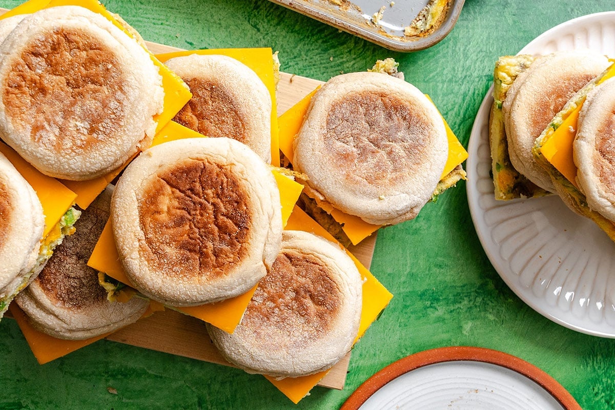 A group of breakfast sandwiches with English muffins, cheddar cheese, and egg, arranged on a cutting board and a plate on a green surface.
