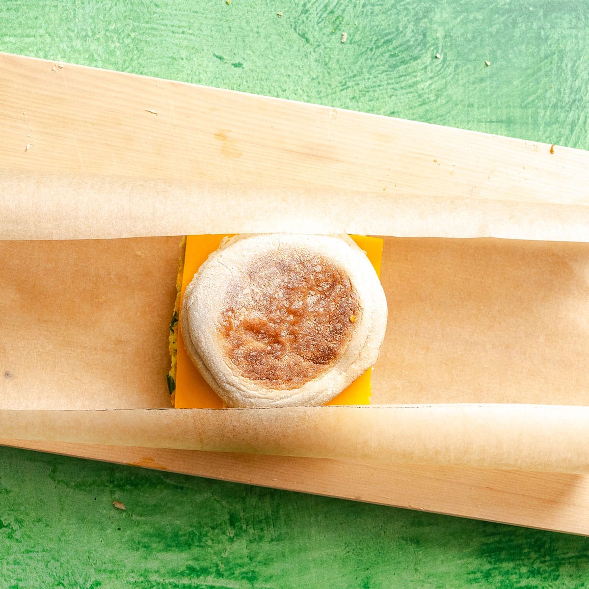 An English muffin breakfast sandwich with cheddar cheese is partially wrapped in parchment paper on a green surface.