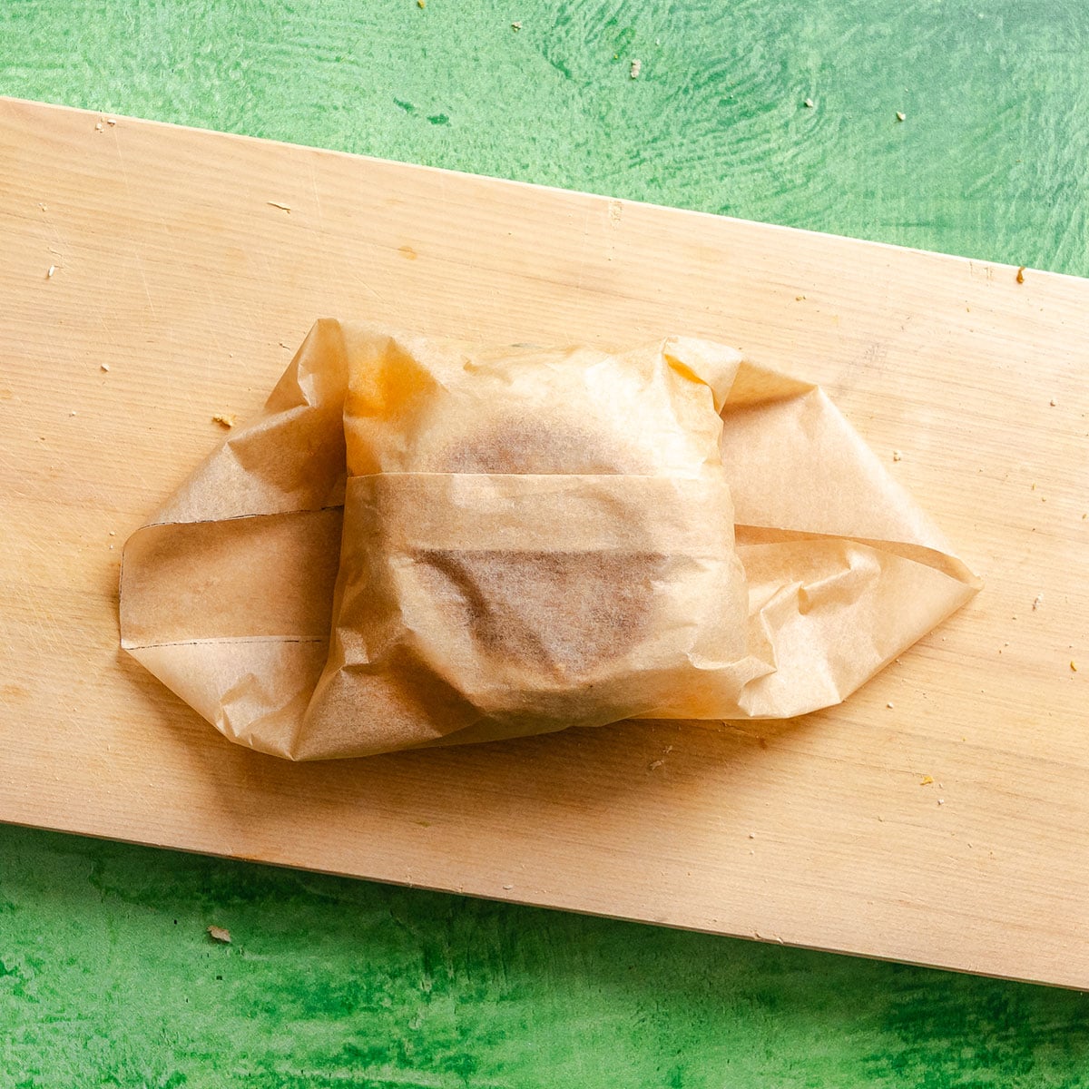 A wrapped sandwich sits on a wooden board, with brown parchment paper partially folded around it against a green background.