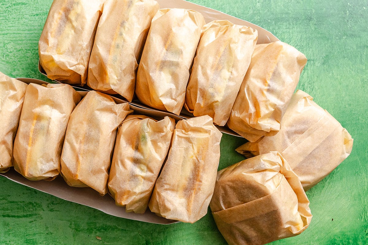 Ten sandwiches wrapped in brown paper are arranged in two trays on a green surface, with two additional wrapped sandwiches placed beside them.