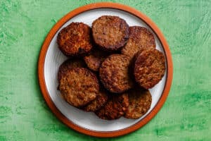 A white plate with a brown rim holds several browned vegan sausage patties, arranged in a pile, on a textured green background.