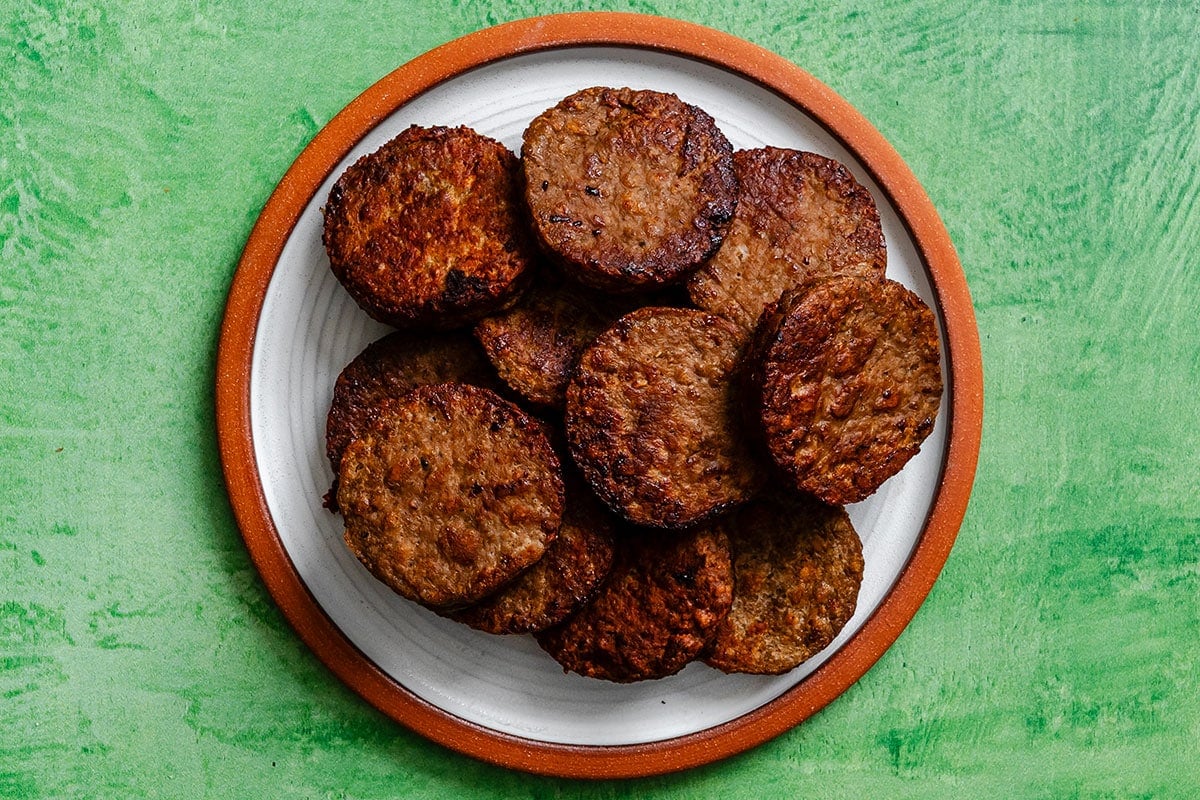A white plate with a brown rim holds several browned vegan sausage patties, arranged in a pile, on a textured green background.