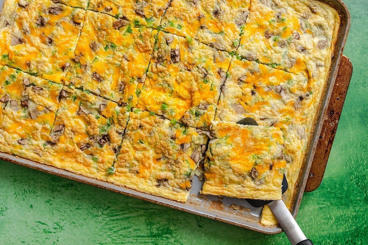 A baked egg casserole with cheese, mushrooms, and herbs cut into square portions on a baking sheet, with a spatula lifting one piece.