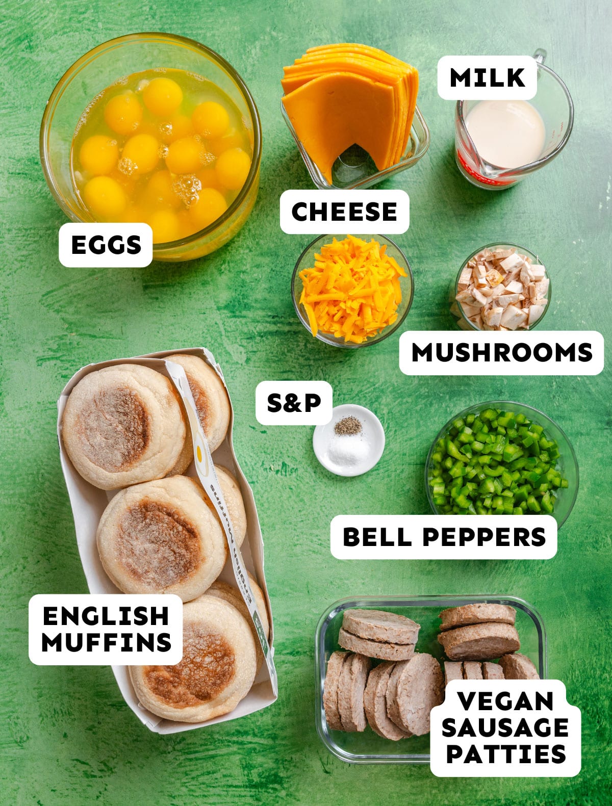Various breakfast ingredients on a green surface, including eggs, milk, cheese, mushrooms, bell peppers, vegan sausage patties, English muffins, and salt & pepper—perfect for assembling vegetarian breakfast sandwiches. Each item is labeled.