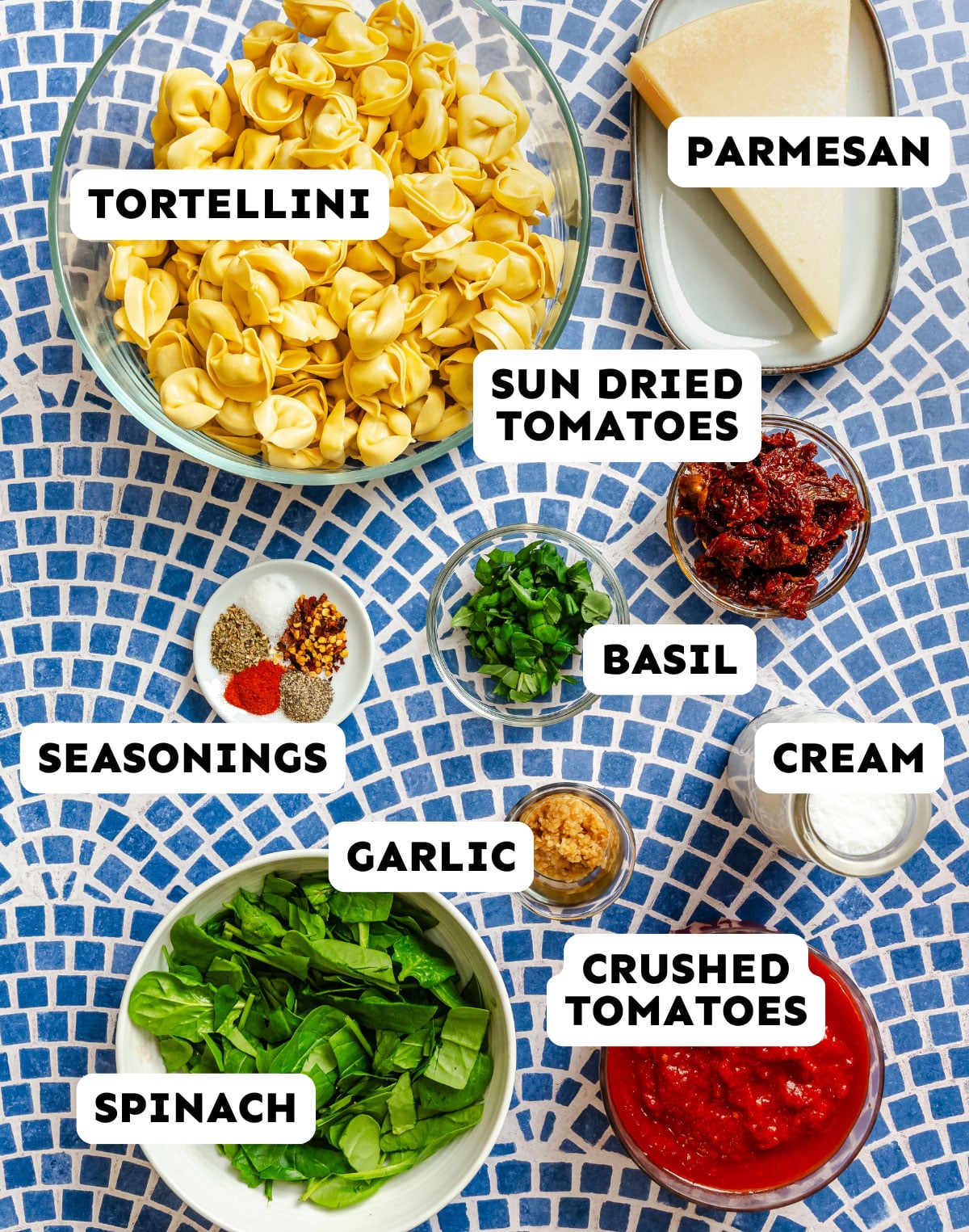 Bowls and containers of labeled ingredients for a pasta dish, including tortellini, parmesan, sun-dried tomatoes, basil, cream, seasonings, garlic, crushed tomatoes, and spinach.