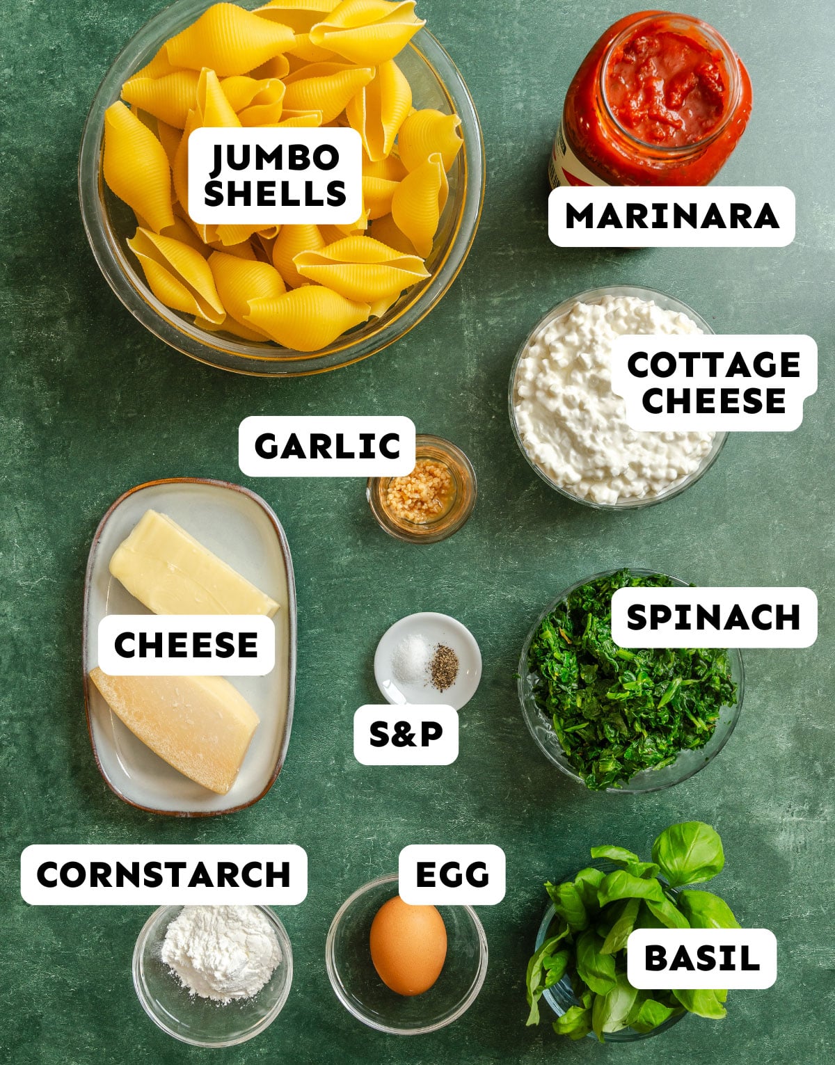 Ingredients for stuffed shells are arranged on a green surface, including jumbo shells, marinara, cottage cheese, spinach, cheese, garlic, salt and pepper, cornstarch, egg, and basil.