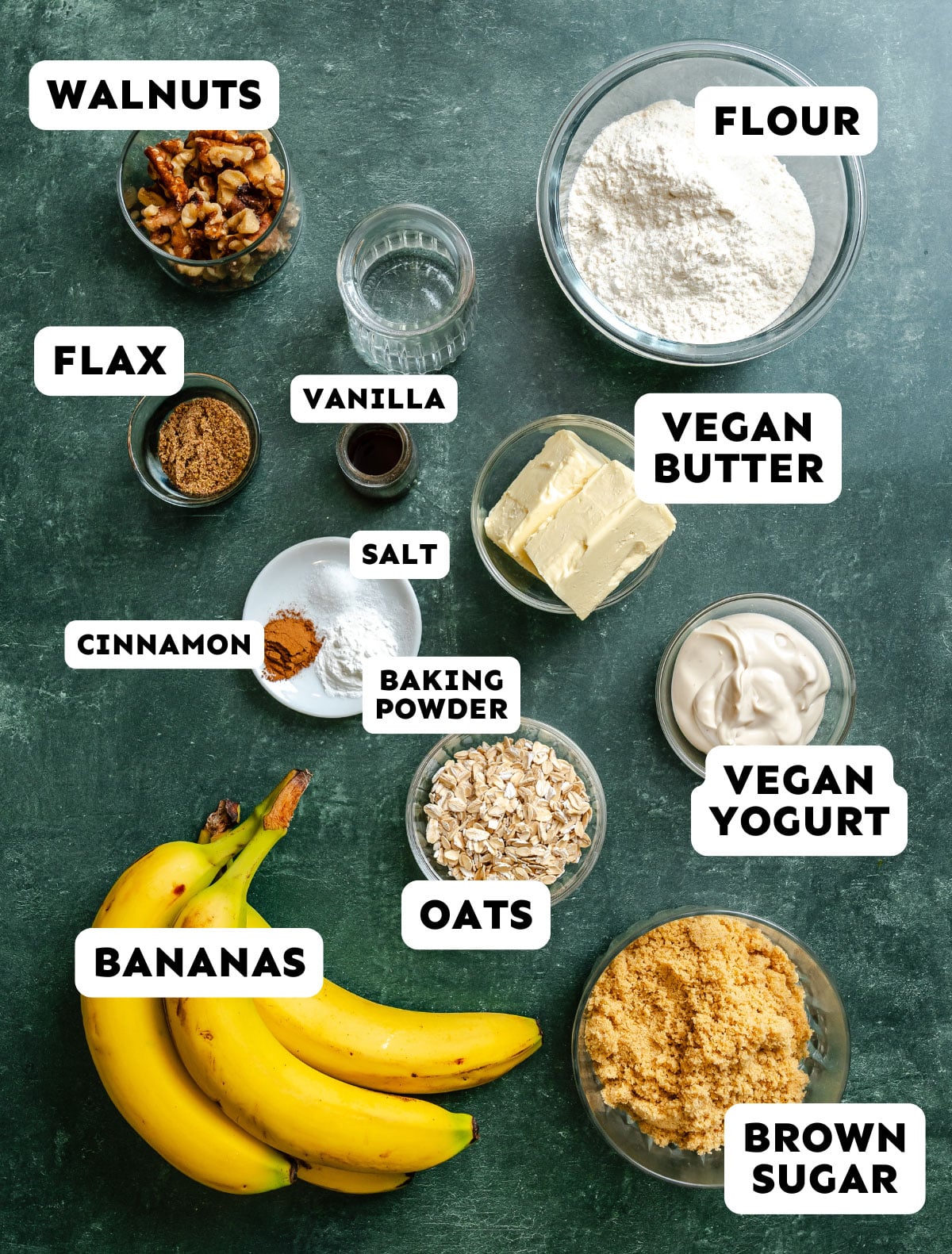 Various labeled baking ingredients including flour, vegan butter, vegan yogurt, brown sugar, bananas, oats, walnuts, flax, vanilla, cinnamon, salt, and baking powder on a dark surface.