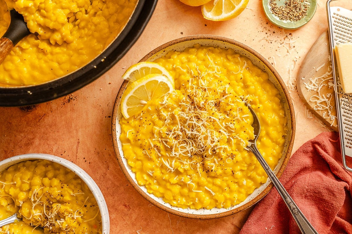 A bowl of creamy yellow lemon pepper Italian penicillin garnished with grated cheese, black pepper, and a lemon wedge, with a spoon, next to a pot, cheese grater, and lemon slices on a wooden surface.