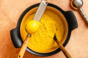 A pot of yellow risotto with a lemon and a grater resting on top, alongside a spoon and a slotted utensil on a brown surface.