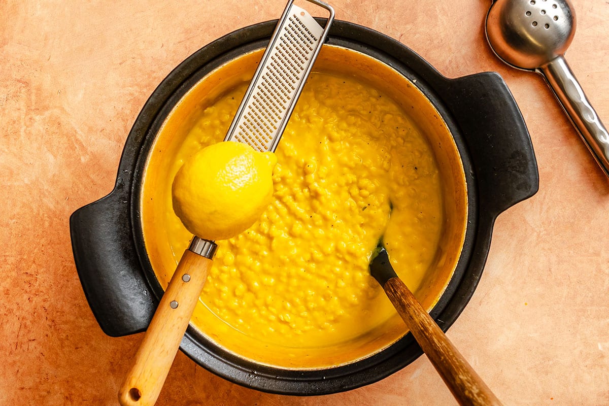 A pot of yellow risotto with a lemon and a grater resting on top, alongside a spoon and a slotted utensil on a brown surface.