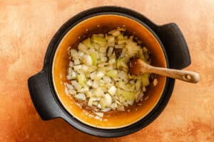 Chopped onions being sautéed in a large black pot with a wooden spoon on an orange surface.