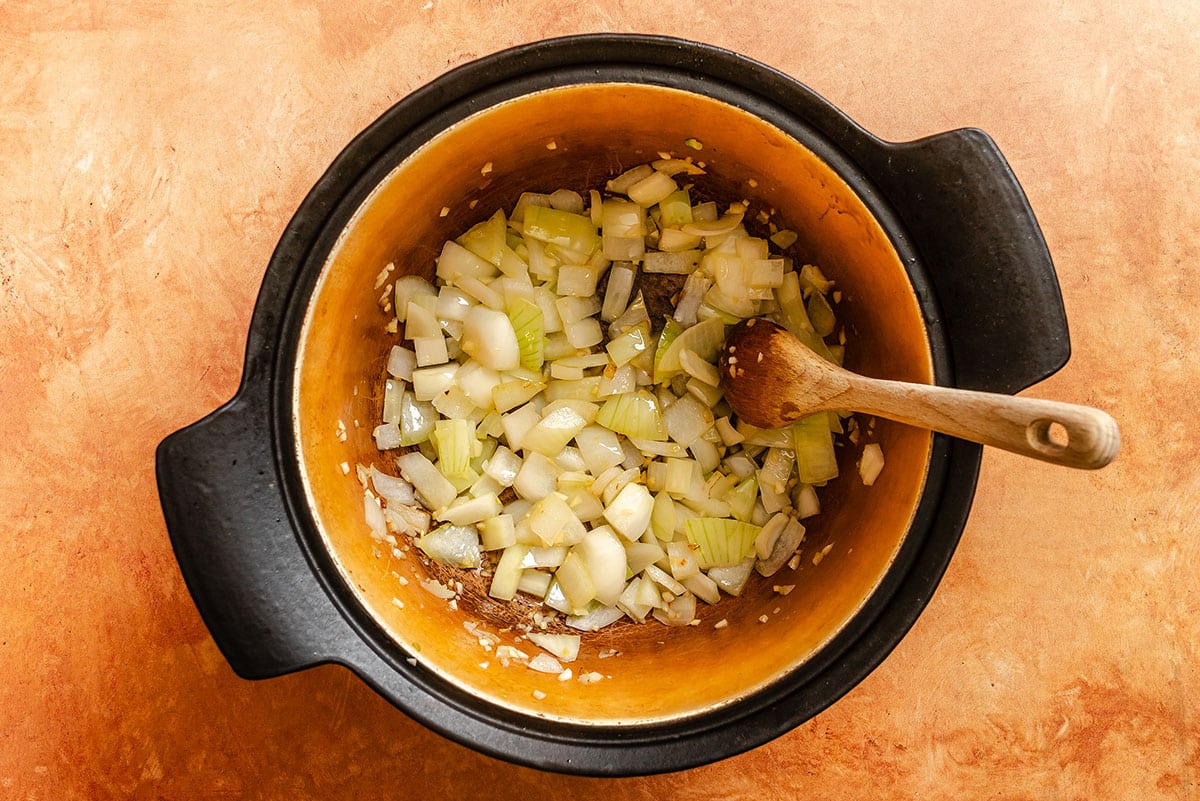 Chopped onions being sautéed in a large black pot with a wooden spoon on an orange surface.