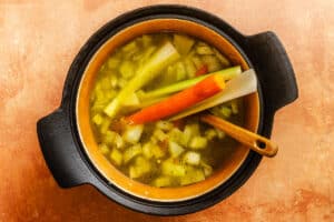 A pot of vegetable soup with chopped onions, carrots, and celery simmering in broth, with a wooden spoon resting inside.