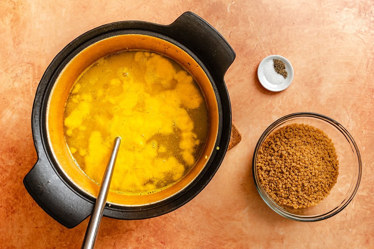 A pot of yellow liquid with a spoon, next to a bowl of brown granules and a small dish of ground pepper on a tan countertop.