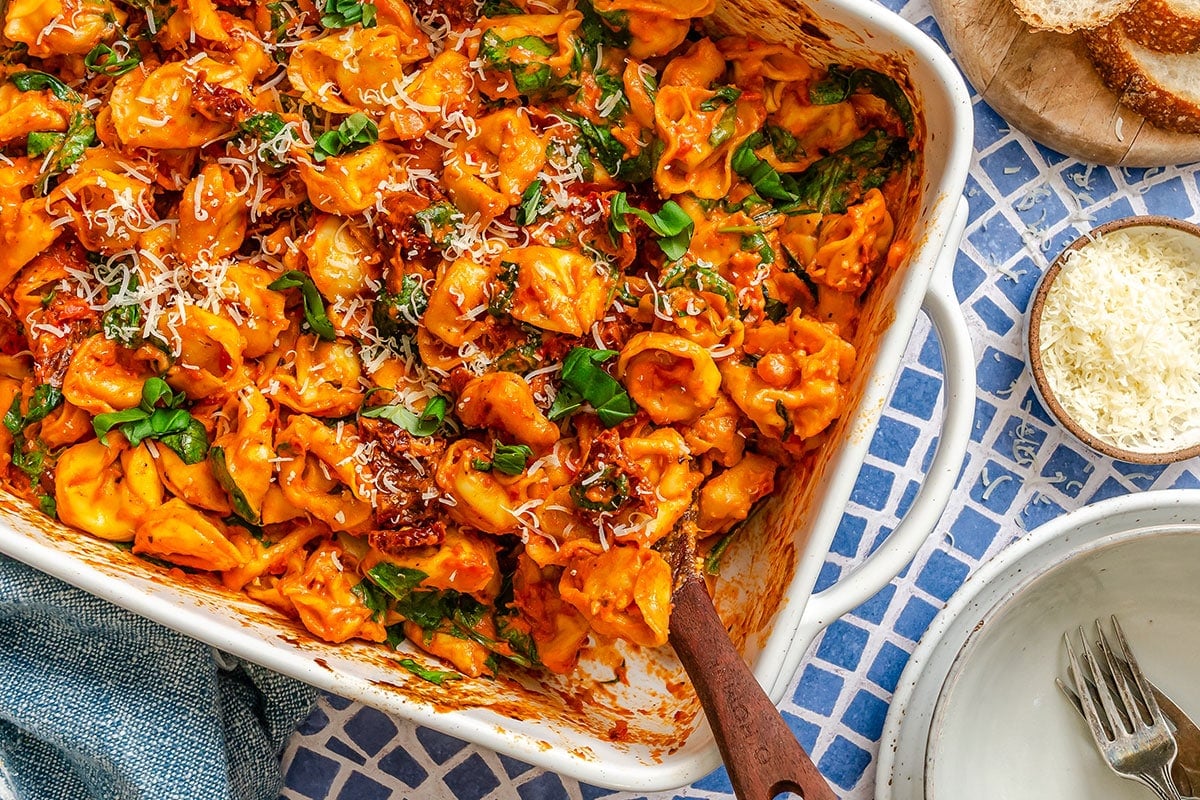 Baked tortellini in tomato sauce topped with grated cheese and fresh basil served in a white dish, with bread and grated cheese on the side.