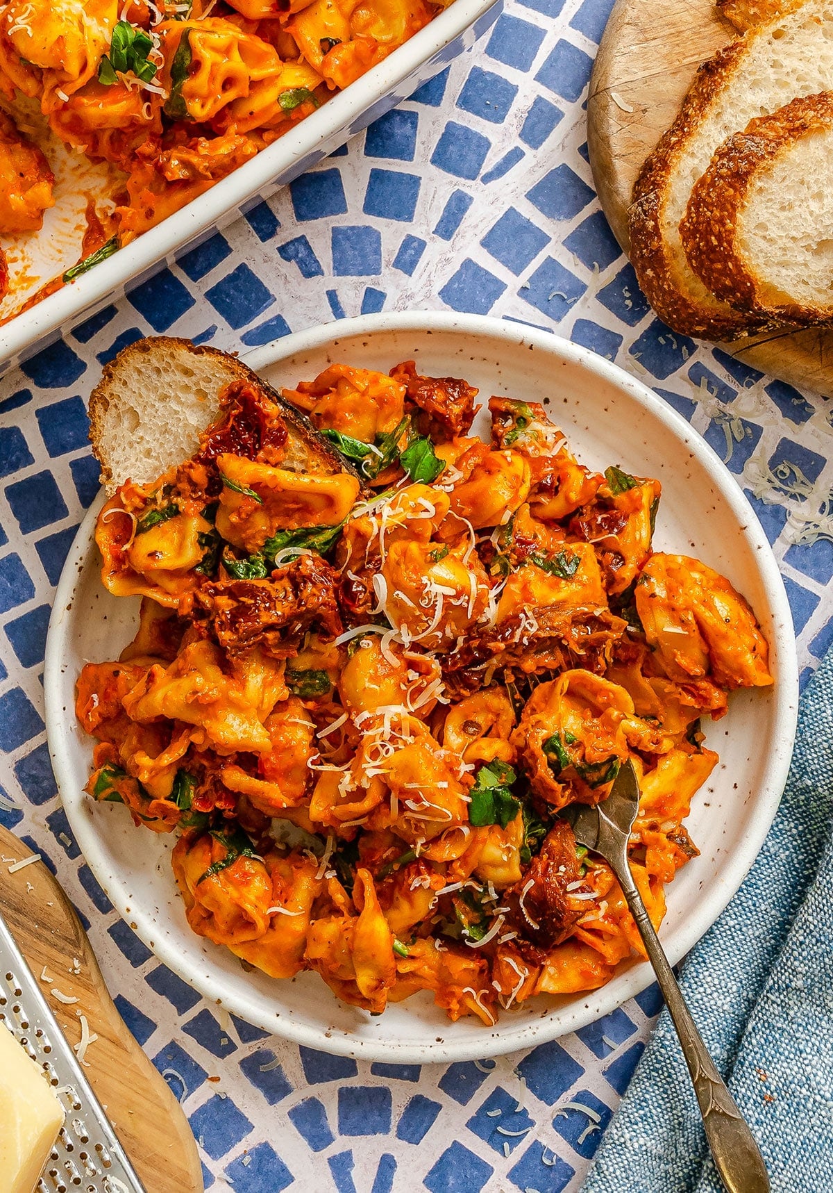 A plate of baked tortellini in tomato sauce topped with grated cheese and herbs, served with a slice of bread on a patterned blue and white table.