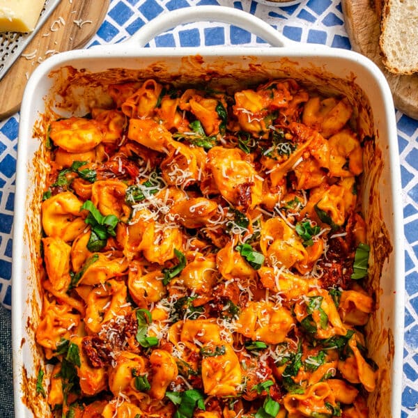 Baked tortellini in tomato sauce topped with grated cheese and fresh basil in a white casserole dish, set on a blue patterned surface.