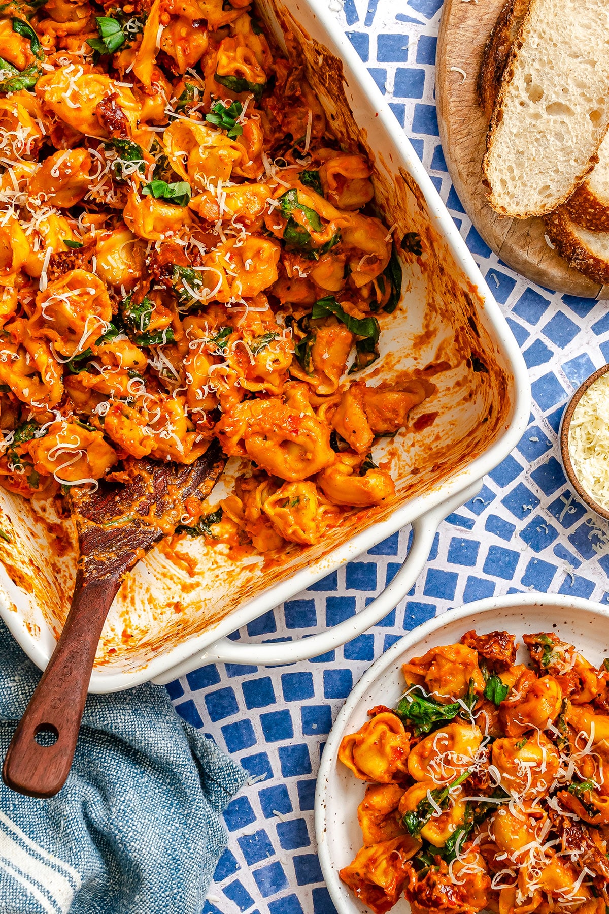A white baking dish filled with baked tortellini in tomato sauce, garnished with basil and grated cheese, sits on a blue patterned surface near a plate of pasta and slices of bread.