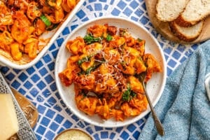 A plate of tortellini pasta in tomato sauce with spinach, topped with grated cheese, served on a blue patterned table with sliced bread and cheese on the side.