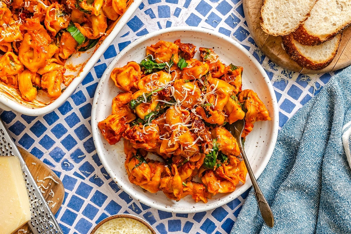A plate of tortellini pasta in tomato sauce with spinach, topped with grated cheese, served on a blue patterned table with sliced bread and cheese on the side.