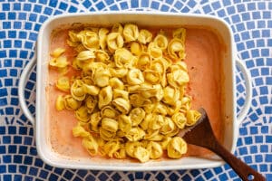 A white baking dish with uncooked tortellini on top of a creamy tomato sauce, placed on a blue mosaic tile surface. A wooden spoon rests in the dish.