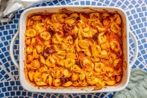 A white baking dish filled with baked tortellini in a red tomato sauce, placed on a blue and white mosaic-patterned surface.