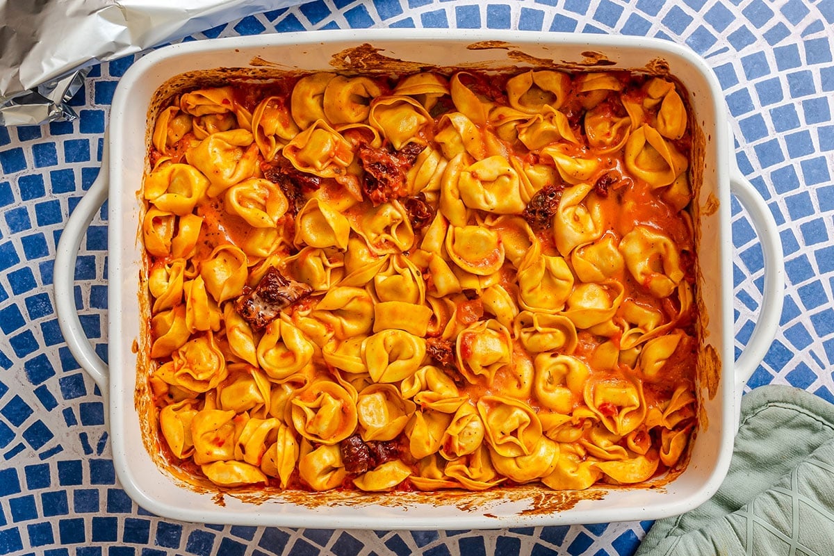 A white baking dish filled with baked tortellini in a red tomato sauce, placed on a blue and white mosaic-patterned surface.