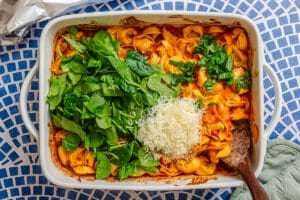 A white baking dish with cooked tortellini in tomato sauce, topped with fresh spinach, basil, and a mound of grated cheese, on a blue and white mosaic surface.