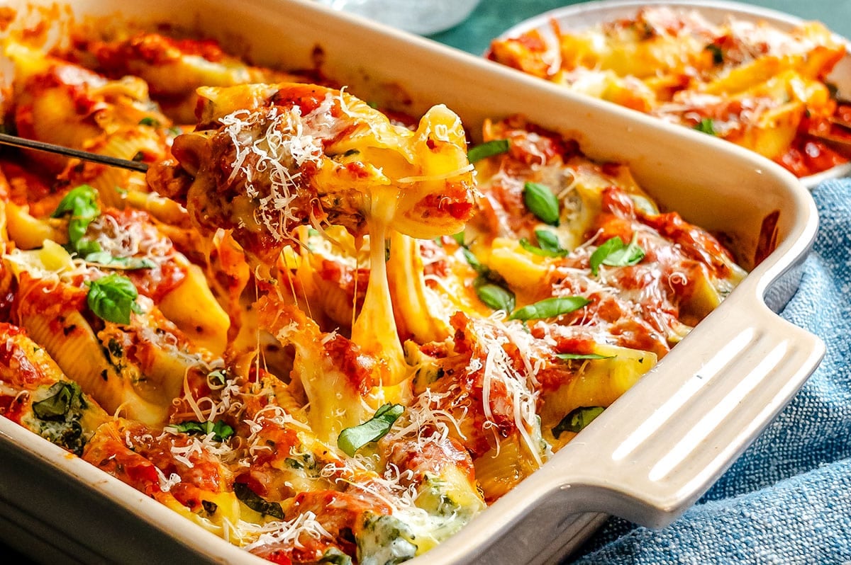 Baked pasta shells stuffed with cheese and herbs, topped with tomato sauce, melted cheese, and basil, being served from a rectangular baking dish.