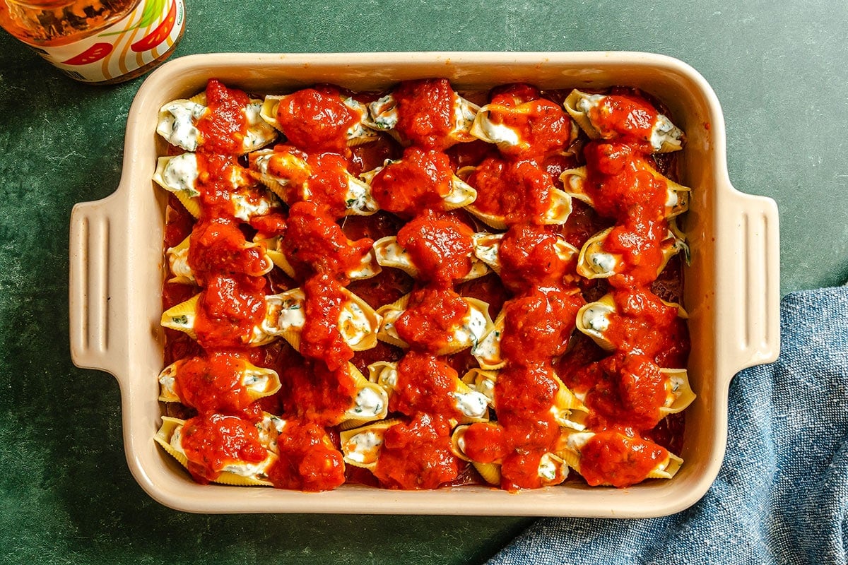 A baking dish filled with pasta shells stuffed with cheese, arranged in rows and topped with marinara sauce, ready to be baked.