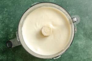 A food processor containing a smooth, creamy white mixture sits on a green countertop.