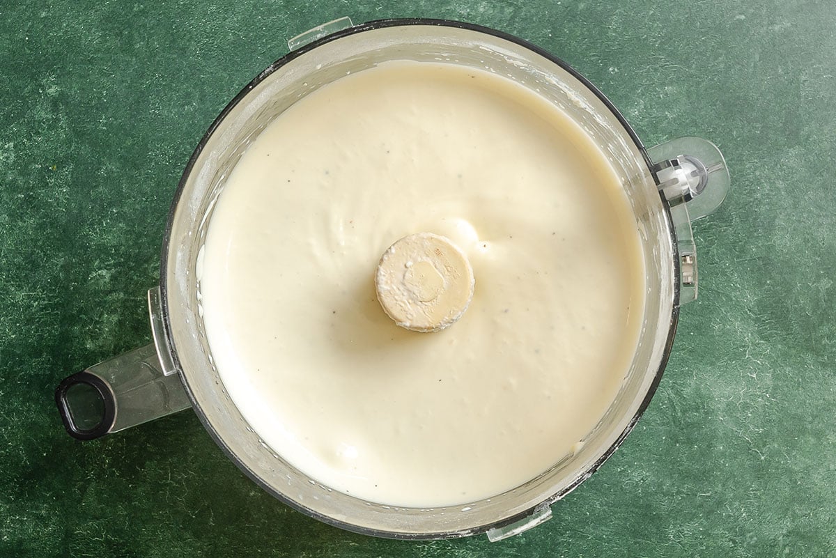 A food processor containing a smooth, creamy white mixture sits on a green countertop.