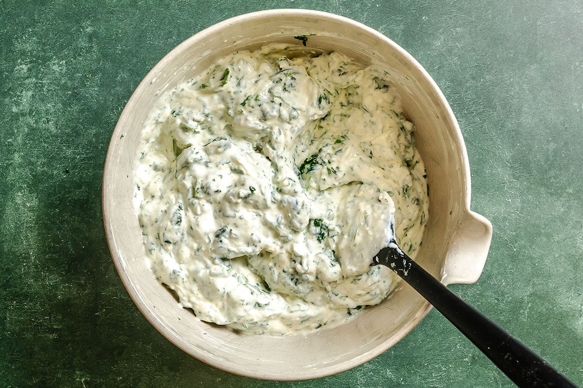 A bowl of creamy spinach dip with visible chopped spinach, stirred with a black-handled spoon, set on a green textured surface.