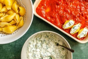Cooked pasta shells, a bowl of whipped cottage cheese and spinach filling, and a baking dish with tomato sauce and stuffed shells are shown on a green surface.