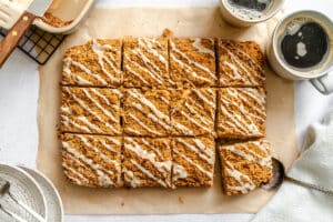 Rectangular iced coffee cake cut into 12 squares on parchment paper, with two cups of coffee, plates, and a serving utensil nearby.