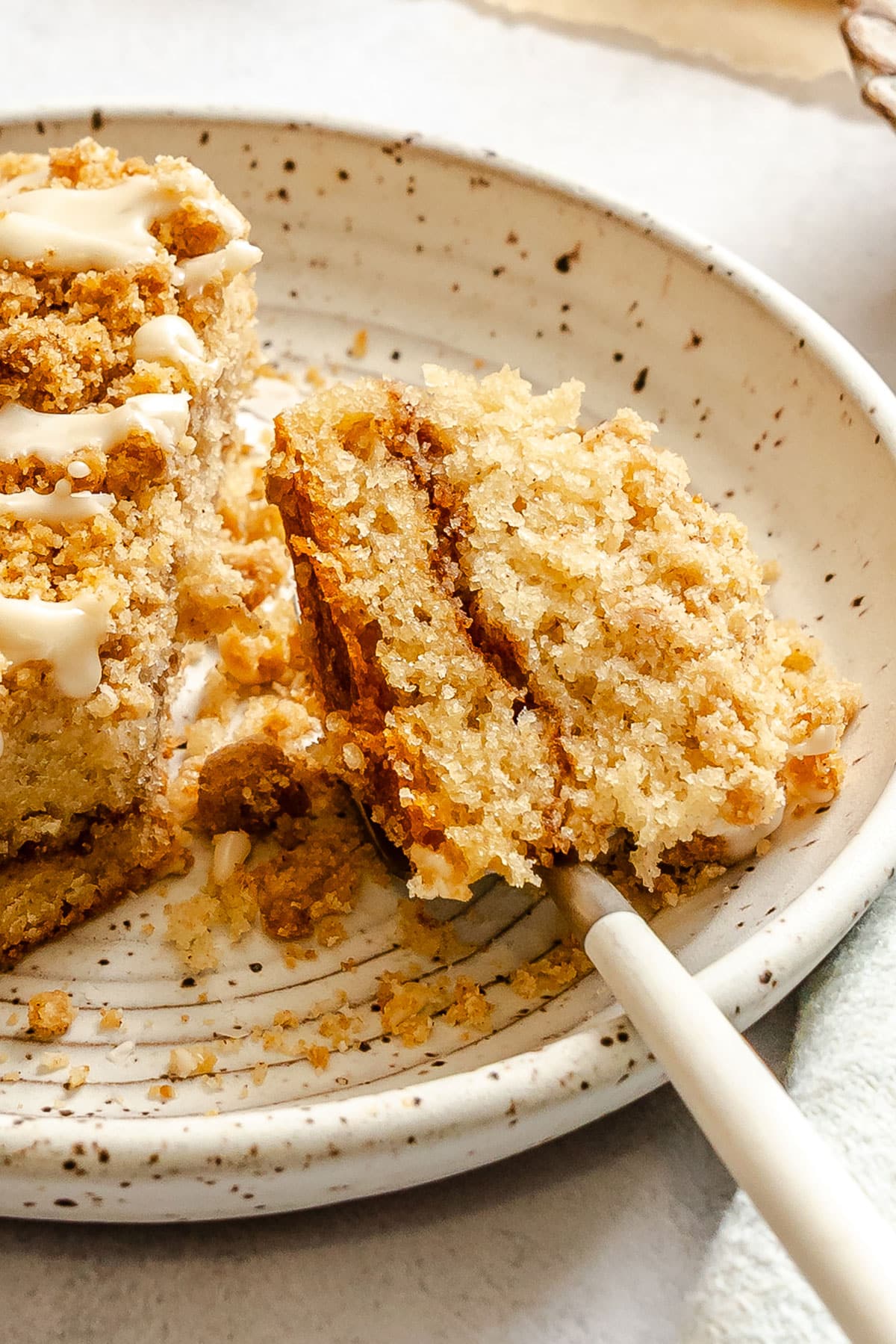 A fork holds a bite of crumbly coffee cake with icing, resting on a speckled ceramic plate beside the remaining slice.